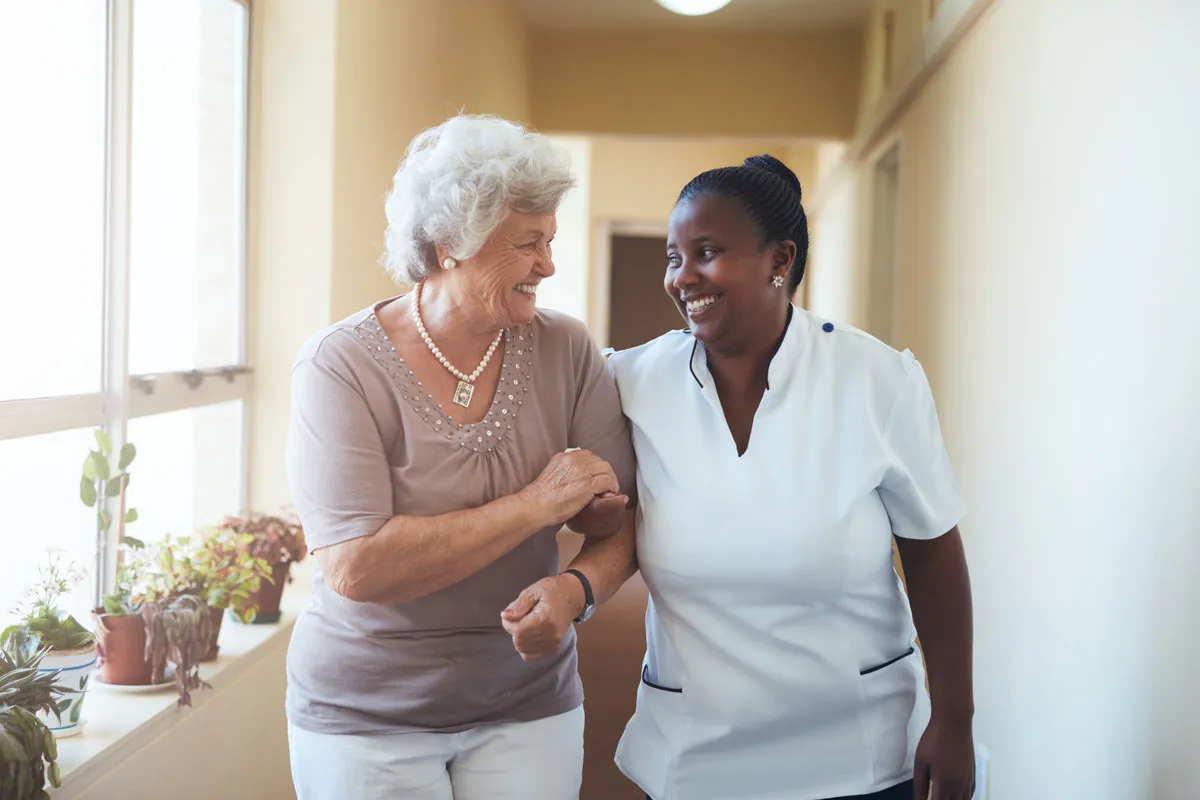 A caregiver walking arm in arm with an older woman down a sunlit hallway.