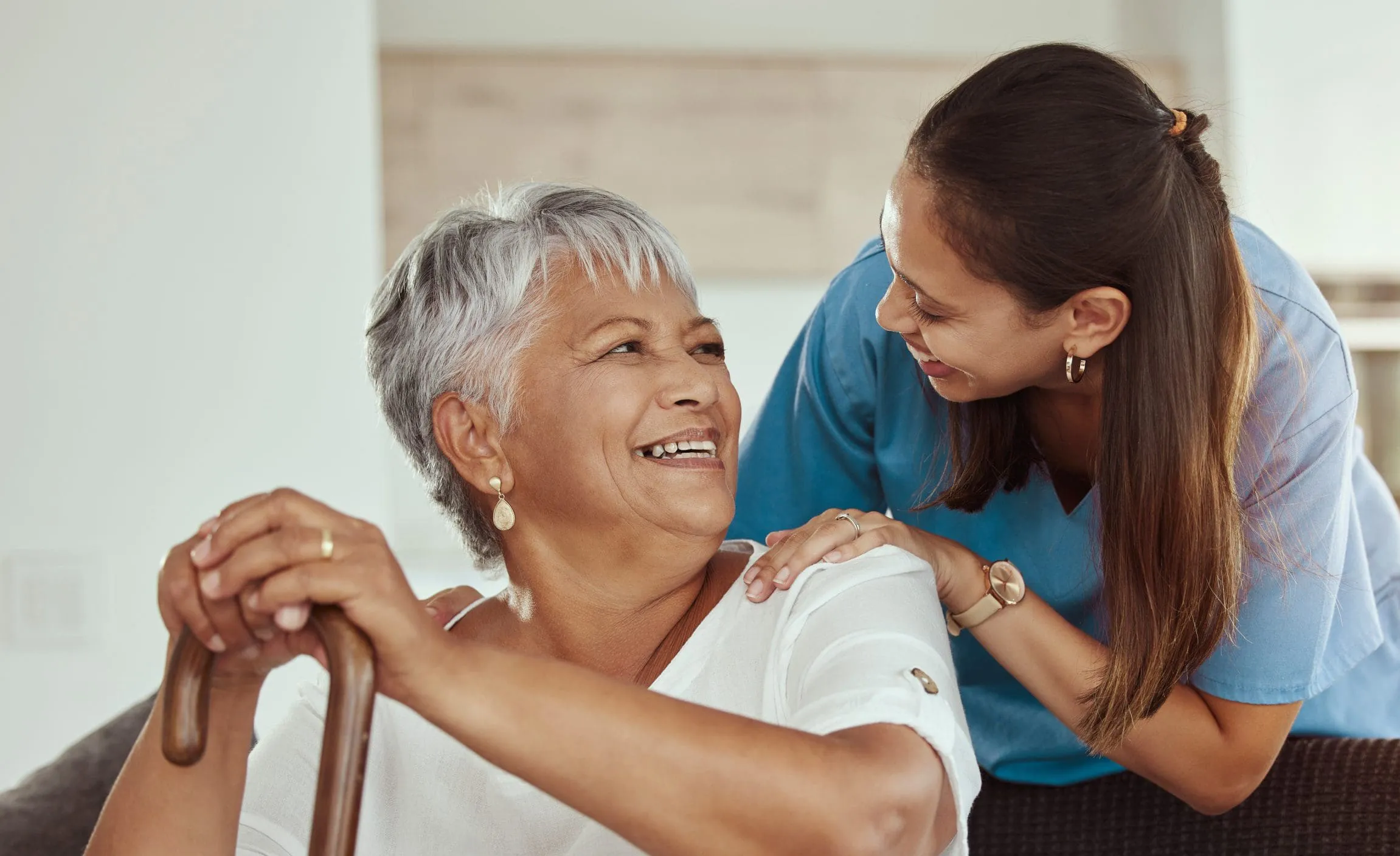 A smiling caregiver with her hand on the shoulder of an older woman holding a cane.