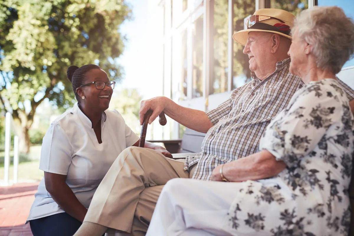 A smiling caregiver kneels to talk with an older couple seated on a sunny porch.