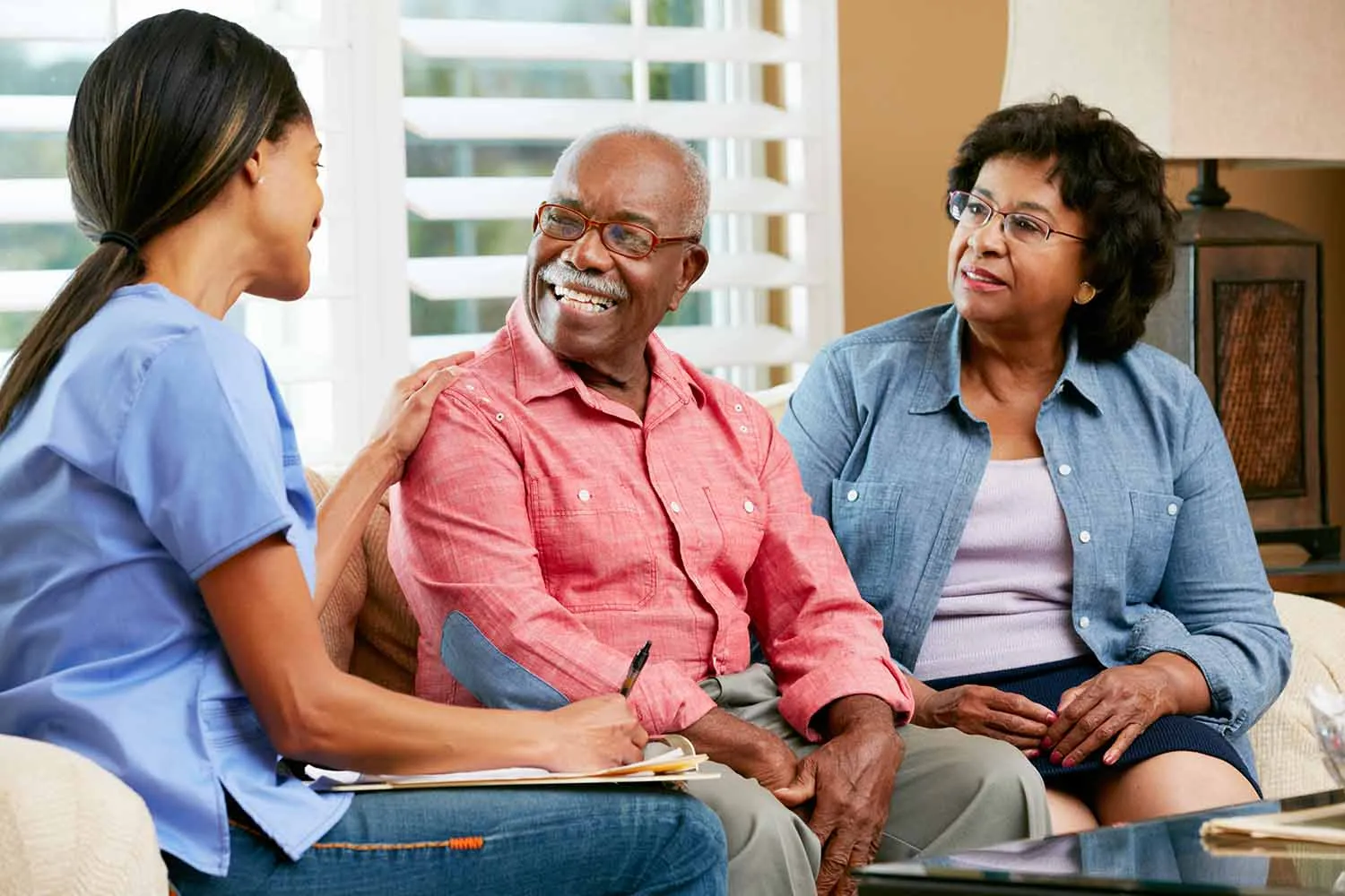 A caregiver in scrubs laughing with an older couple in their living room.