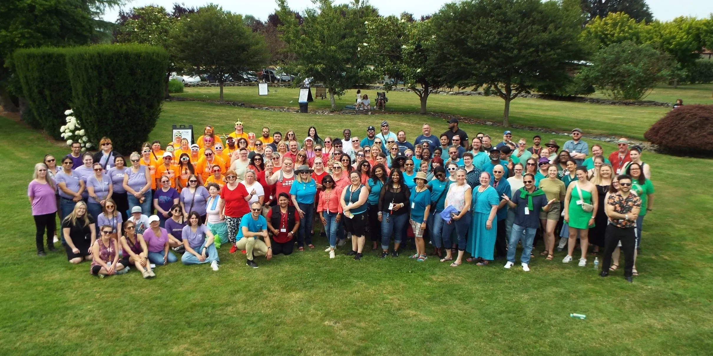 More than a hundred Sunrise Services employees gathered on a lawn for a group photo, many in brightly colored shirts.